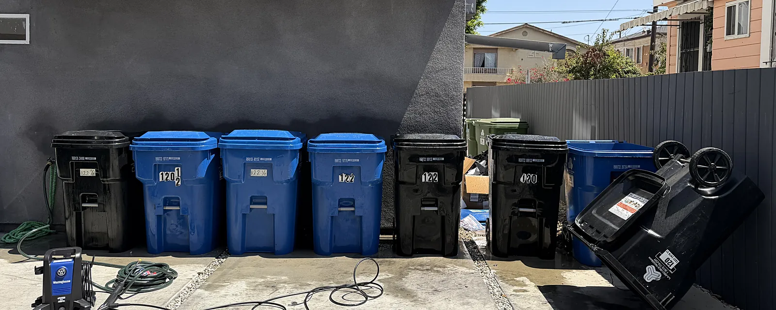 A row of trash and recycling bins lined up for cleaning beside the BrightBin pressure washer and hoses